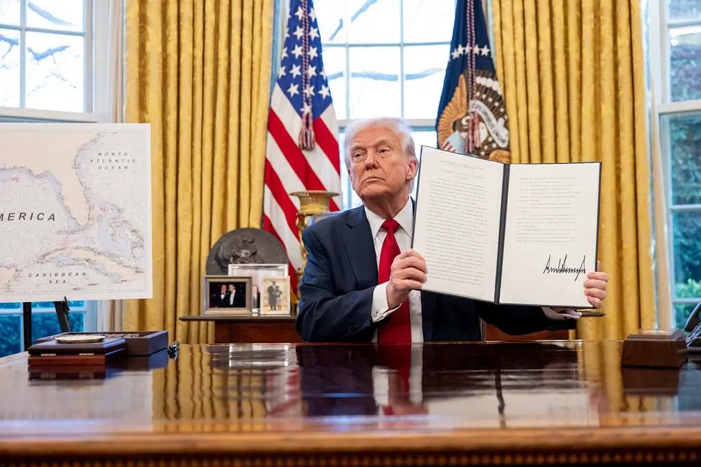 הנשיא דונלד טראמפ יושב במשרד הסגלגל ומציג צו ביצועי חתום להטלת מכסים על יבוא רכב, מרץ 2025, עם דגלי ארצות הברית ודגל הנשיא ברקע